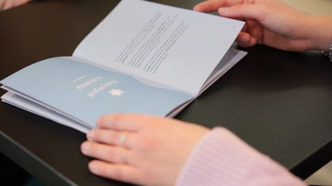 Een opengeslagen boekje 'AUT je hoofd', met op de linkerpagina nog leesbaar 'Positieve gedachten'. De linkerpagina is lichtblauw met witte letters en de rechterpagina lijkt iets lichter blauw met donkere letters. Het boekje ligt op een zwart tafelblad en wordt links opengehouden door een hand met om de ringvinger en middelvinger een ring en rechts is een hand te zien die lijkt te bladeren door het boekje.