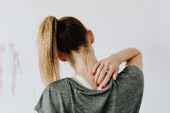 Een blonde vrouw met hoge paardenstaart, vanaf de schouders in beeld, voelt met haar rechterhand net onder haar nek. Ze heeft een gemêleerd grijs shirt met korte mouwen aan en is te zien tegen een lichtgrijze achtergrond met links silhouetten van anatomische weergaven van mensenlichamen.